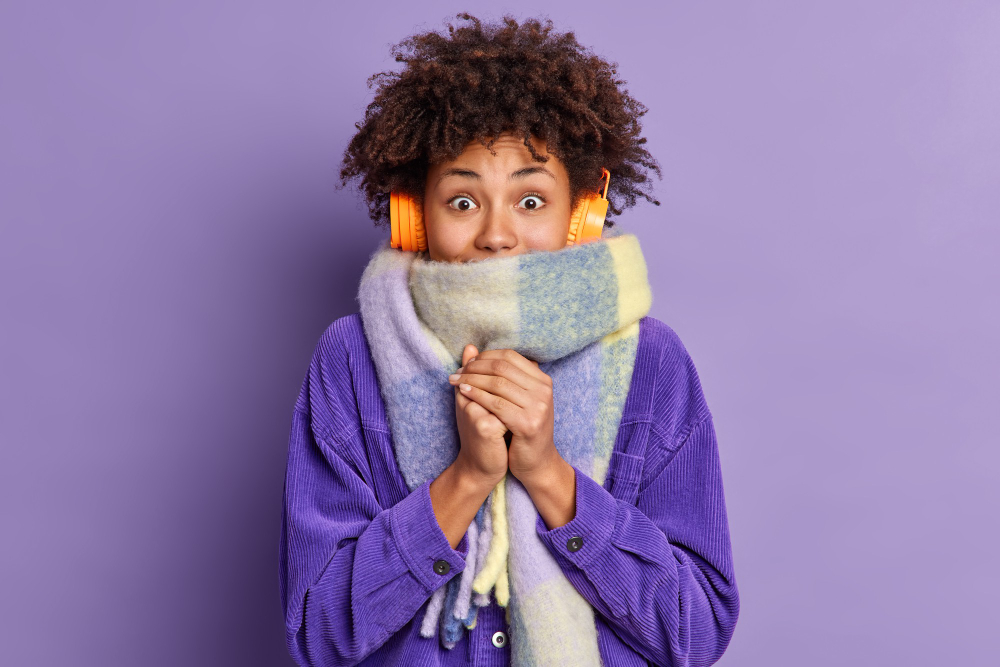 afro american woman feels very cold during freezing weather wears purple jacket warm scarf around neck wals street during winter listens music via wireless headphones keeps hands together